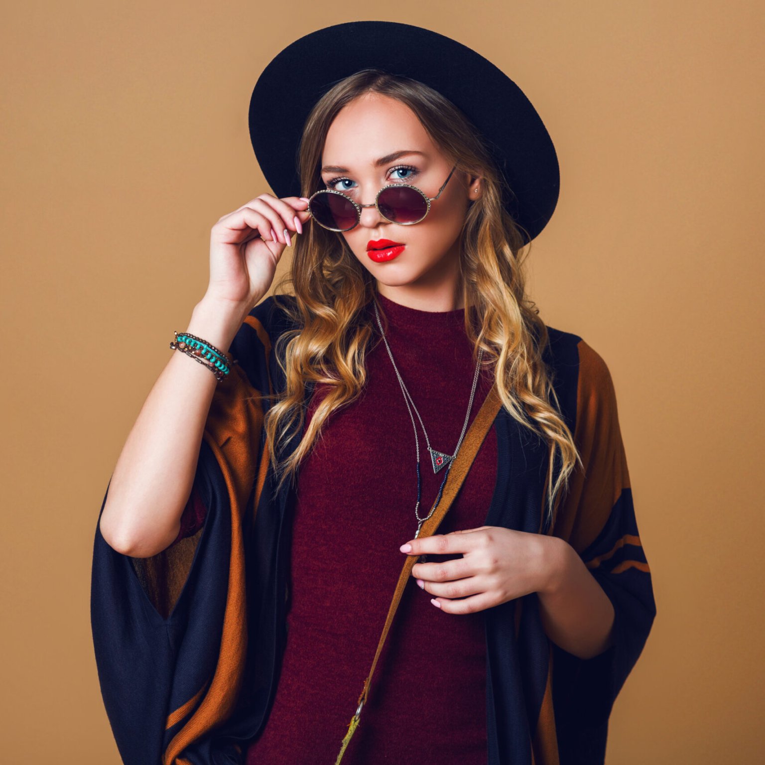 Studio close up portrait of young fresh blonde  woman in brown straw poncho , wool black trendy hat and round glasses looking at camera. Green leather had bag.