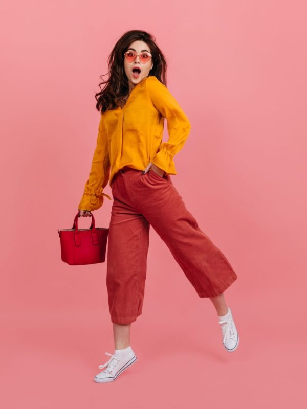 Perky girl in stylish glasses stares into camera in amazement, walking on pink background. Brunette in culottes and orange blouse posing with red handbag